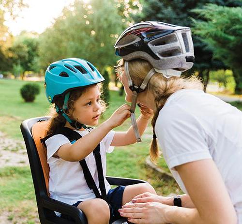 photo of a child in seat on back of a bicycle who is helping their mom fastern her helmet